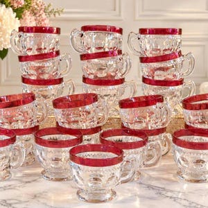 May include: A collection of clear glass cups featuring a textured, dimpled surface and a prominent deep red band around the upper rim. Each cup is designed with a sturdy handle and a short pedestal base, arranged in a multi-tiered display.