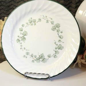 May include: White ceramic plate with a green rim and a delicate green and gold ivy leaf design. The plate has a spiral edge and is displayed on a silver metal stand. A classic piece for serving food.