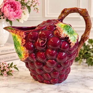 Può includere: Una brocca in ceramica a forma di grappolo d'uva viola scuro con un manico marrone che ricorda un tralcio di vite. La brocca presenta una finitura lucida, un dettaglio a foglia verde ben visibile nella parte superiore e un ampio beccuccio.