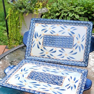 Könnte beinhalten: Zwei rechteckige Serviertabletts aus Keramik mit Griffen, die eine weiße Mitte mit blauen Blumenmustern und gelben Akzenten aufweisen, umgeben von einem blau-weißen geometrischen Rand. Die Tabletts haben eine glänzende Oberfläche.