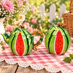 Può includere: Una teiera in ceramica e una zuccheriera coordinata con coperchio, entrambe a forma di anguria con buccia a strisce verdi, centro rosso brillante con semi neri e manici e coperchi a forma di viticcio marrone su una tovaglia a quadri.
