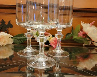 May include: Three clear, stemmed wine glasses with a decorative base and a wide bowl. The glasses are arranged on a reflective surface, with flowers in the background. The glasses are made of clear glass.