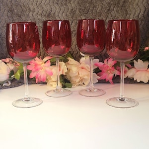 Pode incluir: Quatro taças de vinho tinto rubi com hastes e bases transparentes. As taças estão dispostas em fila, mostrando o seu design elegante. O fundo apresenta flores e uma superfície cinzenta texturizada.