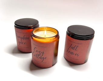 May include: Three amber glass candle jars with labels. The candles are labeled "Pumpkin Spice", "Cozy Cottage", and "Fall Leaves".
