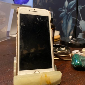 May include: A white marble phone stand with a gold iPhone in it. The phone is on a wooden table with a green stone and glasses next to it.