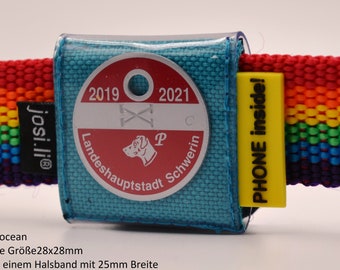 May include: A dog collar with a rainbow-colored strap and a teal blue tag holder. The tag features a red and white emblem with the text "Landeshauptstadt Schwerin" and the years 2019 and 2021. A yellow tag with "PHONE inside!" is attached.