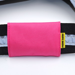 May include: A close-up of a black strap with reflective strips and a bright pink phone holder. The phone holder has a yellow tag that reads "PHONE Inside!"