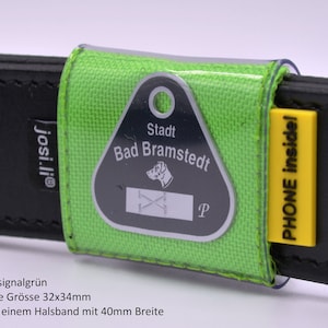 Könnte beinhalten: Ein grüner Hundeanhänger-Halter mit einer durchsichtigen Plastikabdeckung. Der Anhänger ist silberfarben und trägt die Aufschrift "Stadt Bad Bramstedt" mit einer Hundesilhouette und einem Buchstaben "P". Der Halter ist an einem schwarzen Lederhalsband mit einem gelben Etikett befestigt, auf dem "PHONE inside!" steht.