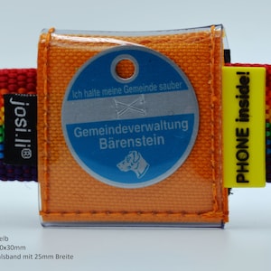 Op de afbeelding: Oranje en gele hondenpenning met een doorzichtige plastic hoes. De penning heeft de tekst "Ich halte meine Gemeinde sauber" en "Gemeindeverwaltung Bärenstein". De penning is bevestigd aan een regenboogkleurig hondenhalsband met de tekst "josi.li®" en "PHONE inside!" op de halsband.