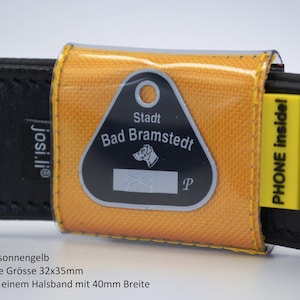 Può includere: Un porta-medaglietta per cani in nylon giallo con una medaglietta nera e argentata che recita "Stadt Bad Bramstedt". Il porta-medaglietta è attaccato a un collare in pelle nera con il testo "josi.li®" e "PHONE inside!" stampato su di esso. La medaglietta per cani misura 32 mm x 35 mm ed è progettata per essere attaccata a un collare largo 40 mm.