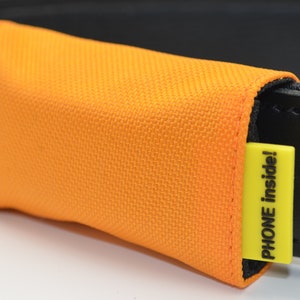 Può includere: Una custodia per telefono arancione brillante attaccata a una cintura nera con un'etichetta gialla che dice "PHONE inside!"