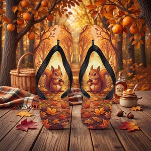Könnte beinhalten: Ein Paar Flip-Flops mit einer gedruckten Illustration eines braunen Eichhörnchens, das eine Eichel in einem herbstlichen Wald mit gefallenen Blättern hält, kombiniert mit schwarzen Gummiriemen vor einem Hintergrund aus Bäumen.