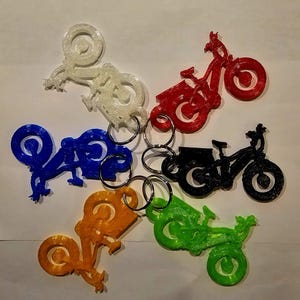 May include: Six colorful keychains shaped like bicycles. The keychains are made of plastic and have a shiny finish. The colors are white, blue, red, black, orange, and green.