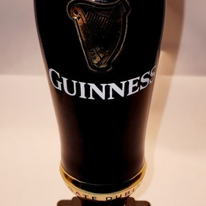 May include: A black Guinness beer glass with a white foam top and the Guinness logo. The glass is on a black base with the words "Gate Dublin" in gold lettering.