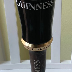 Puede incluir: Un tirador de cerveza Guinness negro con una parte superior de espuma blanca, un logotipo de arpa dorado y el texto "ESTD 1778 GUINNESS" y "ST. JAMES'S GATE DUBLIN". El tirador est&aacute; unido a una base de madera con la palabra "GUINNESS" impresa en ella.