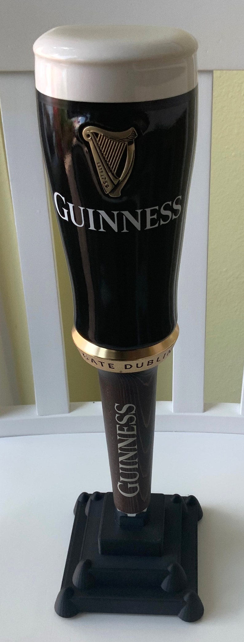 Puede incluir: Un tirador de cerveza Guinness negro con una parte superior de espuma blanca, un logotipo de arpa dorado y el texto "ESTD 1778 GUINNESS" y "ST. JAMES'S GATE DUBLIN". El tirador est&aacute; unido a una base de madera con la palabra "GUINNESS" impresa en ella.