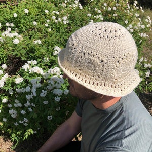 May include: A crocheted hat in a cream color with a wide brim. The hat has a textured design with small holes throughout. The hat is worn by a person sitting in a garden.