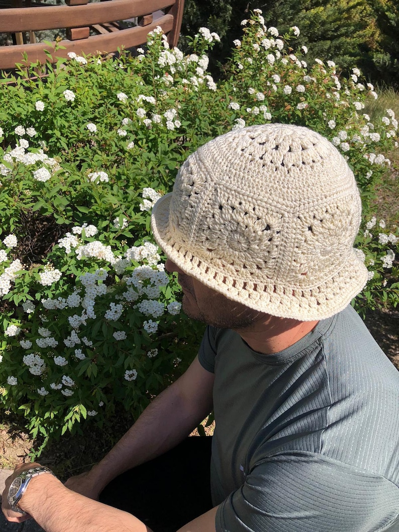 May include: A crocheted hat in a cream color with a wide brim. The hat has a textured design with small holes throughout. The hat is worn by a person sitting in a garden.