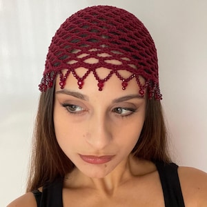 Puede incluir: Un cubrecabezas de ganchillo burdeos con un diseño similar a una red. El cubrecabezas está adornado con cuentas rojas colgantes a lo largo de la frente. El cubrecabezas se lleva sobre cabello oscuro.
