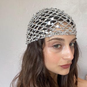 May include: A silver sequined headpiece featuring a crocheted net design. The accessory is constructed from metallic silver yarn woven into an open grid pattern, with small, reflective circular sequins attached to the intersections of the mesh.