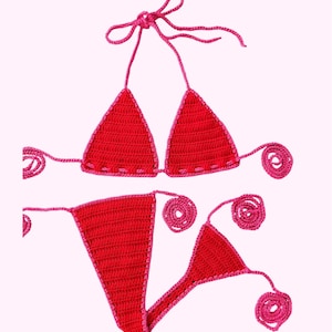 May include: A handmade red crochet bikini set with pink trim. The triangle top has halter straps and the bottoms are a thong style. The edges of the bikini are finished with a contrasting pink color, and the ties have decorative spiral ends.