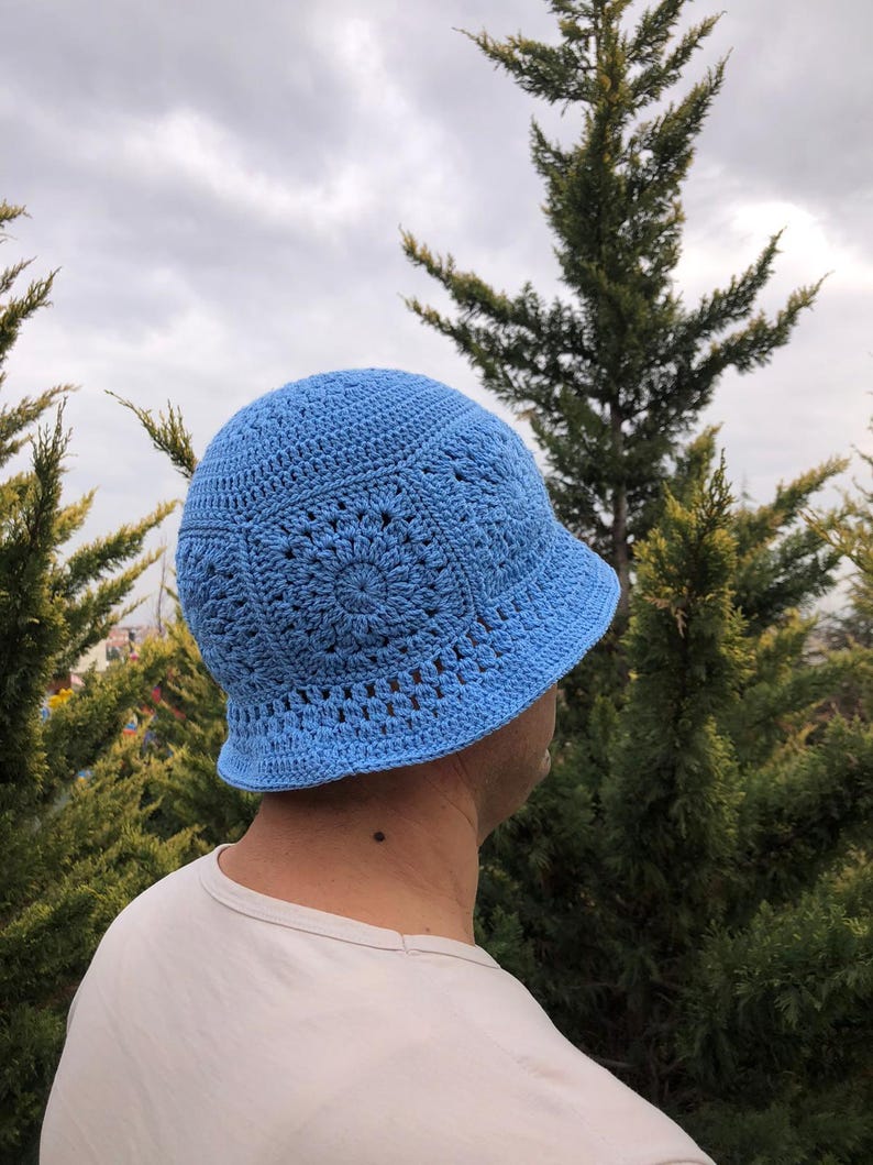 May include: A light blue crocheted bucket hat with a detailed pattern. The hat is worn by a person standing in front of a green tree.