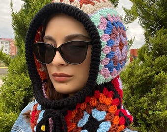 May include: A colorful, crocheted hood with a black trim and ties. The hood features a patchwork design with squares of various colors, including red, orange, blue, and green. The hood is worn with a denim jacket.