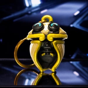 Peut inclure: Un porte-clés représentant un personnage stylisé en forme de gland avec un corps crème, un capuchon doré et des bordures dorées. Le personnage possède de grands yeux circulaires noirs avec des accents bleu vif et un petit détail facial sombre.