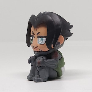 May include: A small figurine of a character with black hair, a goatee, and green eyes. The figure is wearing a gray outfit with a green and white backpack. The character is sitting with their knees pulled up.