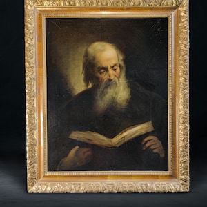 May include: An oil painting of an elderly man with a long white beard, reading a book. The man is dressed in dark clothing and is set against a dark background. The painting is in a gold ornate frame.