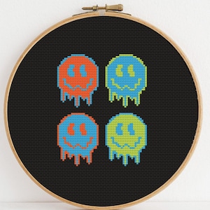 May include: Four pixelated smiley faces with dripping edges in different colors: orange, blue, green, and yellow. The faces are on a black background inside a wooden embroidery hoop.