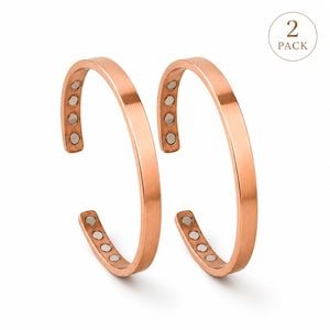 May include: Two copper-colored magnetic bracelets. Each bracelet is a simple, open cuff design with a flat surface. Small, round magnets are embedded in the inner surface of each bracelet. The bracelets are displayed on a white background.