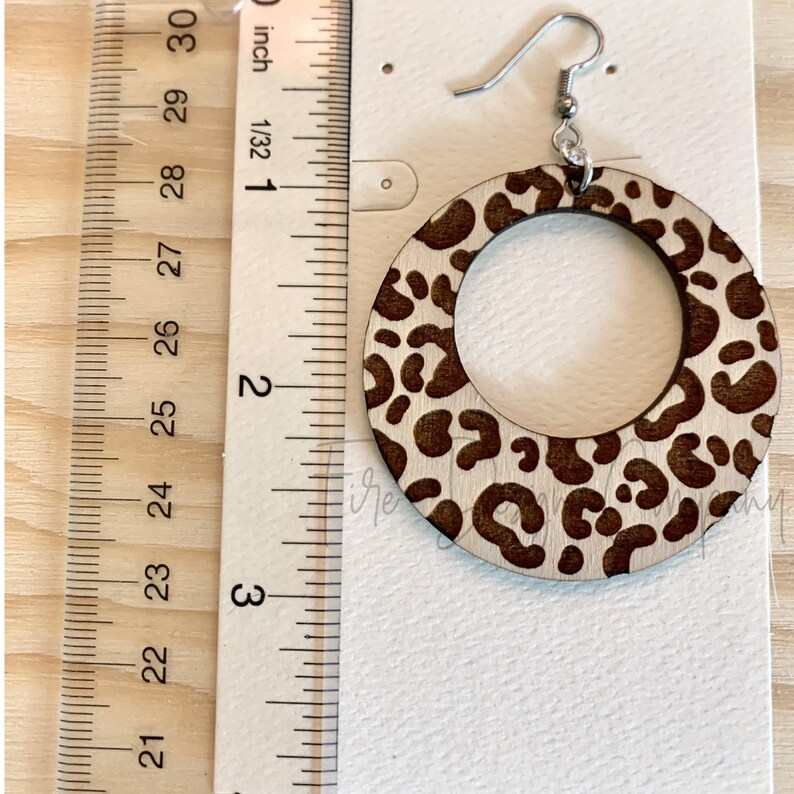 May include: A pair of wooden earrings with a leopard print design. The earrings are approximately 2 inches in diameter.