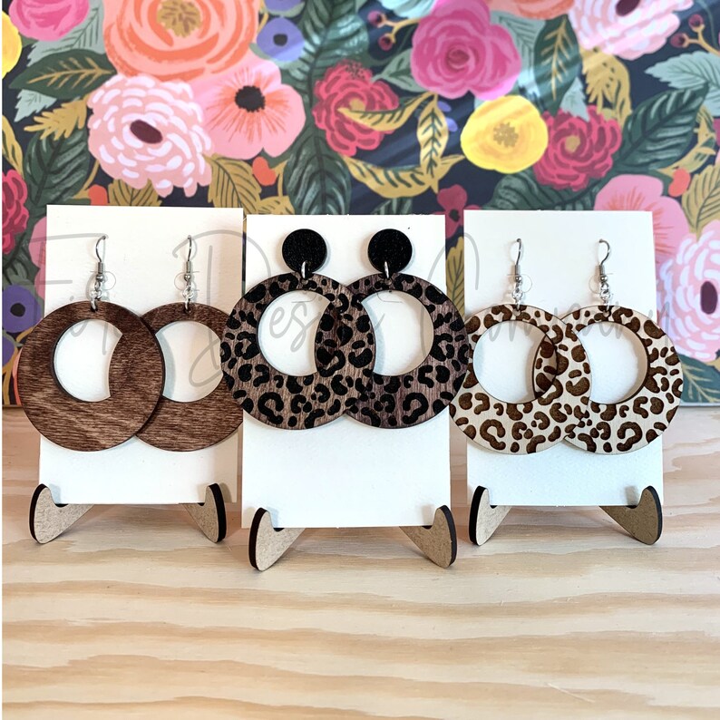 May include: Three pairs of wooden hoop earrings with a leopard print design. The earrings are displayed on white stands.