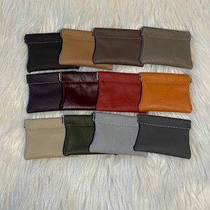 May include: A collection of twelve rectangular leather coin pouches arranged in three rows. Each pouch features a top zipper closure, visible stitching along the upper edge, and a smooth texture, available in various colors including black, tan, brown, gray, purple, burgundy, red, orange, beige, olive green, and slate.