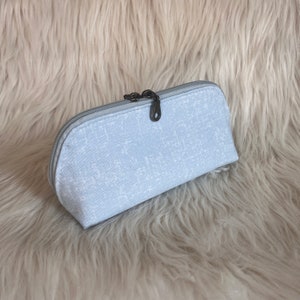 May include: A light gray zippered pouch with a subtle white pattern. The pouch is made of a soft, textured fabric and has a silver zipper pull.