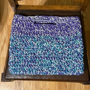 May include: A crocheted tote bag with a purple, teal, and white striped pattern. The bag has a single handle and a small opening at the top.