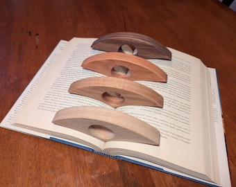 May include: Four wooden thumb page holders in varying shades of brown, resting on the open pages of a book. Each holder has a curved design with a circular opening for the thumb, ideal for reading.