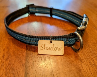 May include: A black and teal woven pet collar featuring a rectangular light-colored wooden tag attached with a small metal ball chain. The tag is engraved with the name Shadow in a clear, dark font. The collar includes a black plastic buckle.