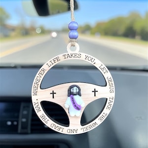 Puede incluir: Amuleto de coche de madera con forma de volante, con el texto "Wherever life takes you, let Jesus take the wheel and lead the way." Presenta una pequeña figura de Jesús y dos cruces. Incluye una cuerda para colgar.