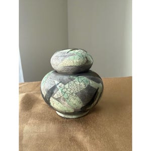 Puede incluir: Un tarro de cerámica con tapa que presenta un acabado de esmalte craquelado en tonos verde menta, gris carbón y negro. El recipiente tiene un cuerpo redondeado y abombado con una tapa a juego, mostrando un patrón texturizado tipo red.