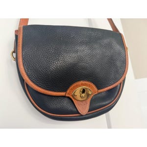 May include: A navy blue pebbled leather saddle bag featuring tan leather trim and a brass turn-lock closure. The front flap displays an embossed leather patch with the text Dooney & Bourke and USA. The bag includes a matching tan leather shoulder strap.