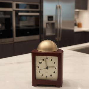 May include: A rectangular dark maroon analog clock with a cream-colored face, black numerals from 1 to 12, and black hands. The clock features a smooth, rounded gold-toned dome on top and sits on four small, rounded feet on a white surface.
