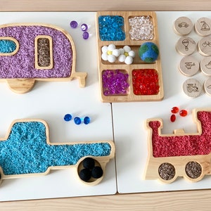 May include: A wooden toy set with three vehicles: a purple camper, a blue truck, and a red train. Each vehicle has a compartment filled with colorful rice. The set also includes a wooden tray with compartments filled with colorful beads and a set of wooden transportation icons.