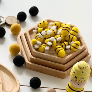 May include: A set of felt bee figurines in yellow and black with white wings, arranged in a wooden hexagonal tray. The tray is on a white surface with other felt balls in yellow and black.