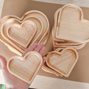 May include: A selection of heart-shaped wooden trays in different sizes. The trays are crafted from light-coloured wood with a smooth, natural finish. Some are stacked, others held in a hand, highlighting their size and shape. These trays are suitable for serving or displaying small items.