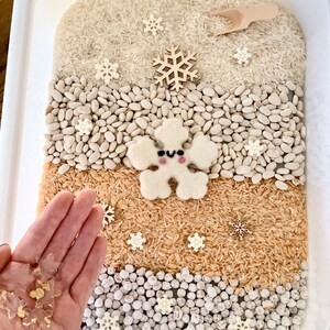 May include: A white plastic container filled with different textures, including white rice, white beans, brown rice, and white beads. There are several small white snowflake decorations scattered throughout the container. A wooden snowflake and a felt snowflake with a smiling face are also visible.