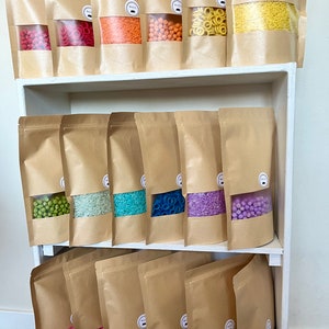 Peut inclure: Une étagère avec plusieurs sacs en papier kraft remplis de perles, anneaux et riz de différentes couleurs. Les sacs sont étiquetés avec un petit logo noir et blanc.