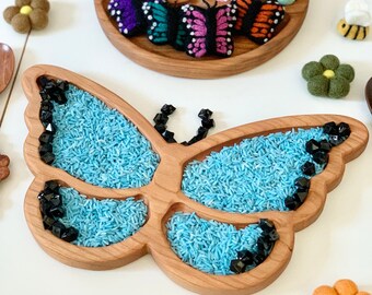 May include: A wooden butterfly-shaped tray with five compartments filled with blue rice and black gems. The tray is on a white surface with a wooden brush and other colorful felt flowers.