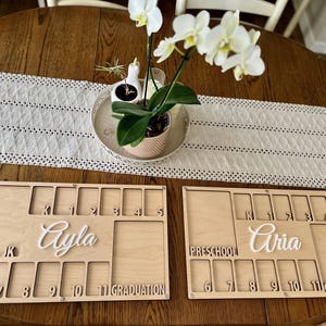 May include: Two wooden milestone boards, each with slots for photos and grade levels from preschool to graduation. The boards feature the names "Ayla" and "Aria" in cursive. A white floral arrangement sits above the boards.